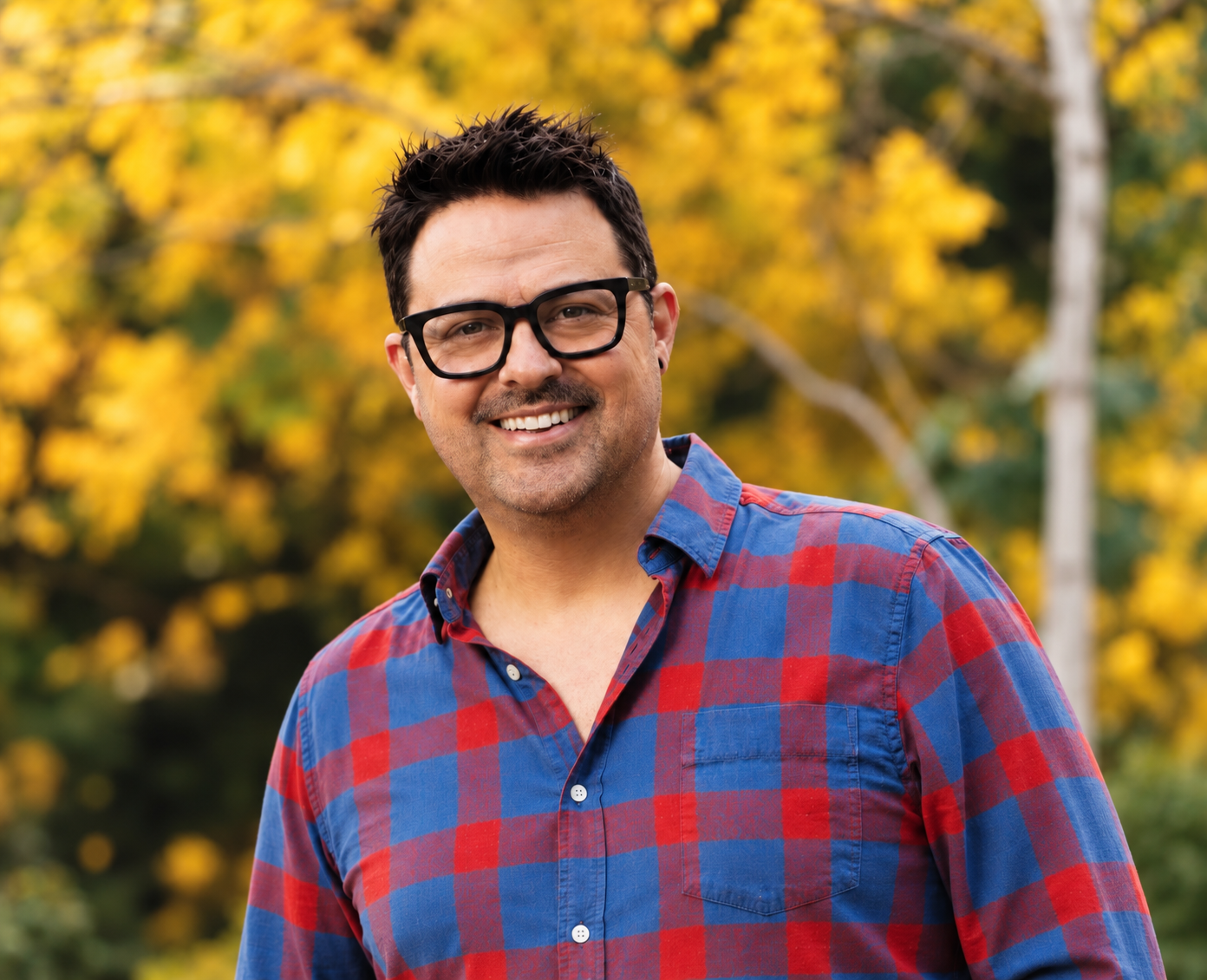 Josh Kostelyk smiling outdoors in front of golden autumn trees.
